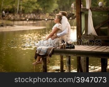 Toned photo of just married couple sitting on pier at river and looking at sunset