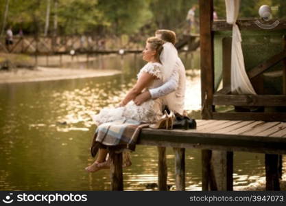 Toned photo of just married couple sitting on pier at river and looking at sunset