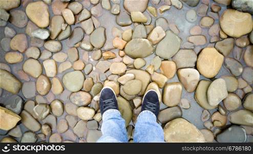 Toned photo of male feet in sneakers standing on pebbles in sea