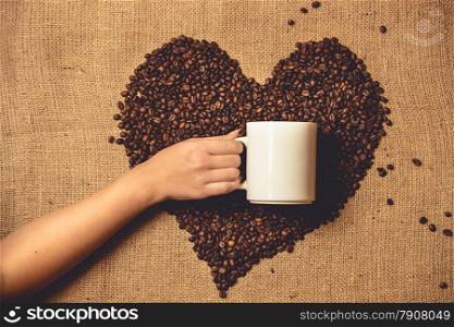 Toned photo of person holding white mug against heart of coffee beans