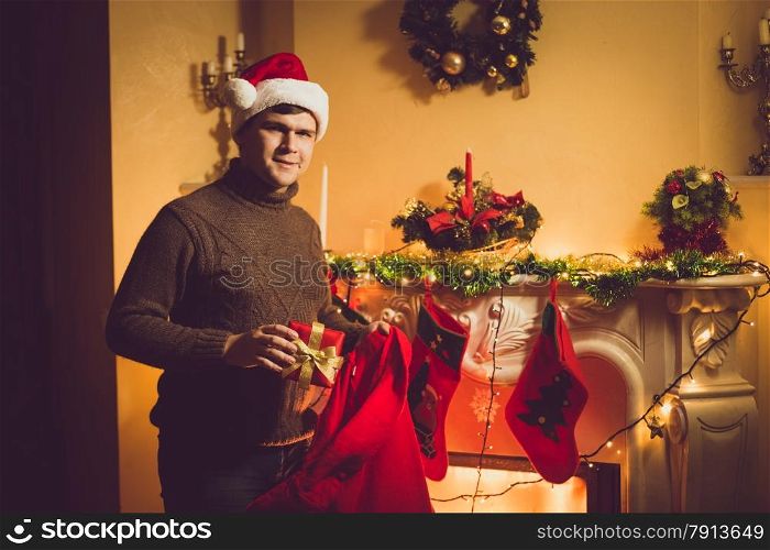 Toned photo of young smiling man taking gift box out of red Santa bag