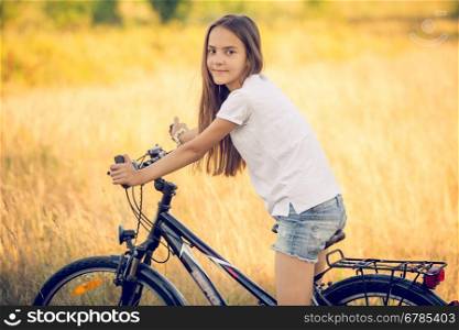 Toned portrait of beautiful teenage girl posing on bicycle at meadow at sunset