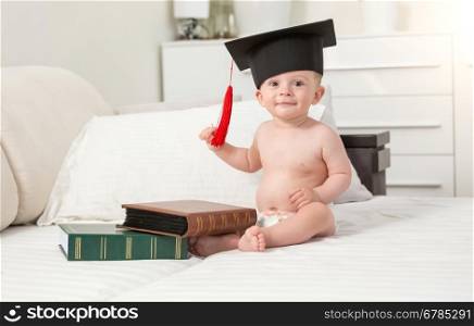 Toned portrait of cheerful baby boy in black graduating cap seating on sofa
