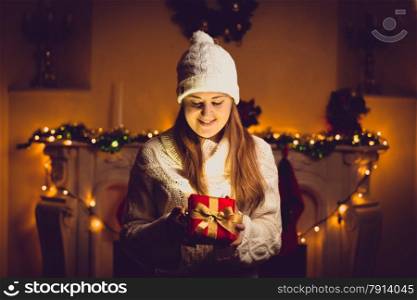 Toned portrait of cute woman in sweater and hat holding glowing gift box at decorated Christmas room