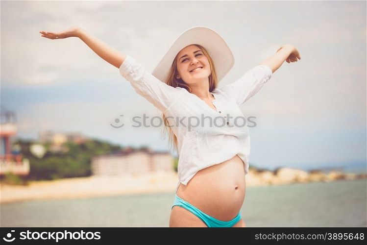 Toned portrait of happy pregnant woman in white shirt enjoying sun on beach