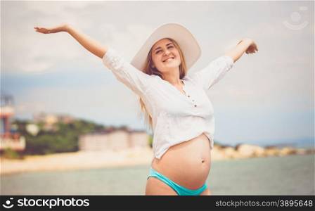 Toned portrait of happy pregnant woman in white shirt enjoying sun on beach