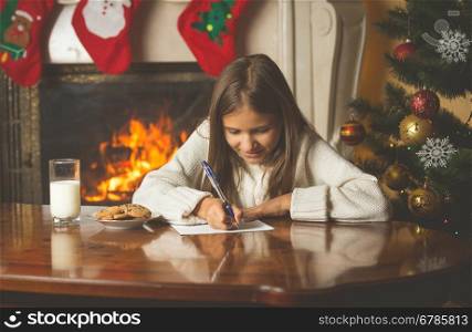 Toned portrait of smiling girl in sweater sitting by the fireplace and writing letter to Santa Claus