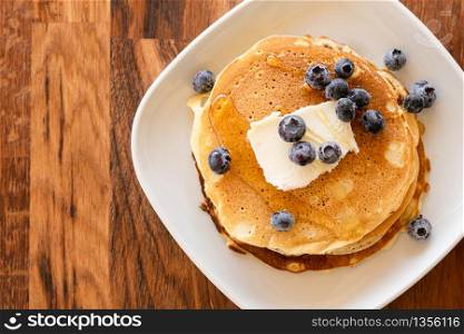 Top angle view on white plate on the wooden board table with pancakes and butter blueberries over and maple syrup or honey
