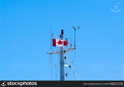 Top of modern metal ship mast against empty blue sky copy space, with antennas, rigging, weather vane, and Canadian flag fluttering in wind.