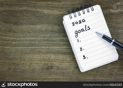 Top view 2020 goals list on notepad. Handwriting on memo paper notebook with black pen over wooden texture background, table blank space. Beginning of new goals and new year resolution concepts.