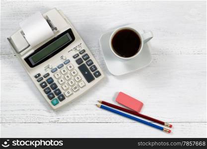 Top view angle of a white wooden desktop with old fashion calculator and paper tape, cup of coffee, pencils and eraser.