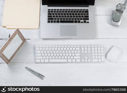 Top view angle of desktop layout for proper use consisting of laptop, keyboard, pens, mouse, picture frame, phone, and work folders on white desk.