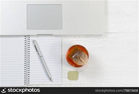 Top view angle shot of desktop with notepad, pen, laptop computer and glass of tea with tea bag inside. Horizontal format.