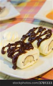 top view crepe rolls filled with white cream and banana, topping with chocolate syrup served with whipping cream on dish