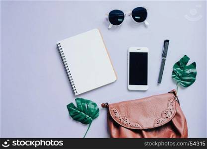 Top view flat lay of woman bag with smartphone, blank notebook, sunglasses and green leaf, copy space