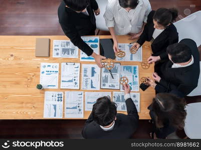 Top view hand holding gear by businesspeople wearing suit for harmony synergy in office workplace concept. Group of people hand making chain of gears into collective form with dashboard report papers.. Top view hand holding gear by group of businesspeople on table in harmony office