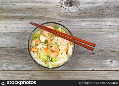 Top view image of Chinese noodle soup in clear glass bowl on top of rustic wood. Layout in horizontal format.