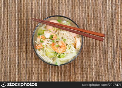 Top view image of Chinese noodle soup in clear glass bowl on top of natural bamboo placemat. Layout in horizontal format.