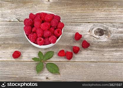 Top view image of fresh Raspberries in white bowl, some falling out, on rustic wood with leaf.