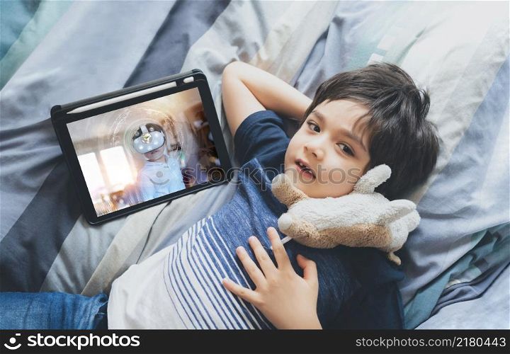 Top view Kid boy using tablet learning online or doing homework, Cute child boy lying in bed with dog toy and looking up at camera with smiling face.