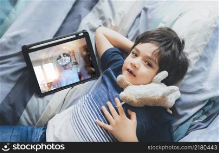 Top view Kid boy using tablet learning online or doing homework, Cute child boy lying in bed with dog toy and looking up at camera with smiling face.