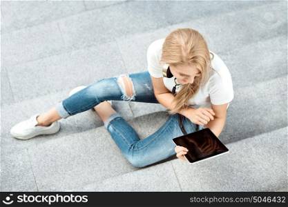 Top view of a beautiful young woman, listening to the music in music headphones, surfing internet on tablet and sitting on stairs.