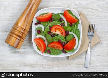 Top view of a freshly made salad, in white plate, with fork on cloth napkin and wooden pepper mill resting on rustic white wood underneath