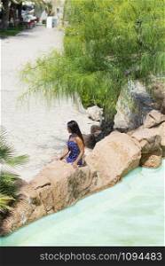 Top view of a pretty young hispanic woman sitting on rock next to a water pond