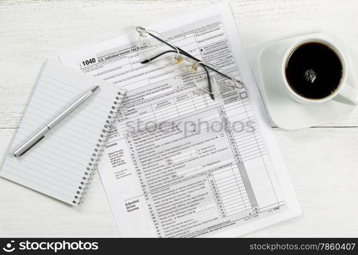 Top view of a silver pen, notepad, tax form, reading glasses and coffee on white desk.