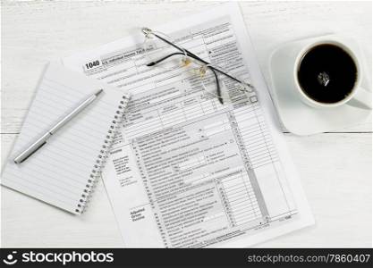 Top view of a silver pen, notepad, tax form, reading glasses and coffee on white desk.