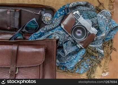 Top view of a vintage photo camera and a brown leather bag with scarf, glasses and pocket watch on sack cloth background. Holiday traveling concept design. Vintage color tone.