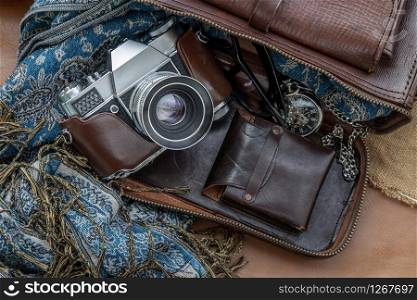Top view of a vintage photo camera and a brown leather bag with scarf, glasses and pocket watch on sack cloth background. Holiday traveling concept design. Vintage color tone.