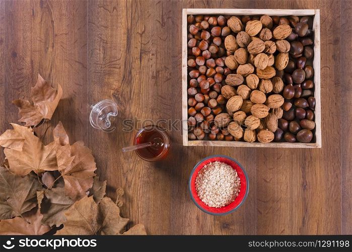 Top view of a wooden box with walnuts, chestnuts and hazelnuts surrounded by a honey jar, a bowl and dry maple leaves on a wooden table. Healthy eating and vegetarian food concept.. Box of nuts with honey jar and dry maple leaves
