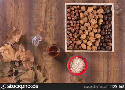 Top view of a wooden box with walnuts, chestnuts and hazelnuts surrounded by a honey jar, a bowl and dry maple leaves on a wooden table. Healthy eating and vegetarian food concept.. Box of nuts with honey jar and dry maple leaves
