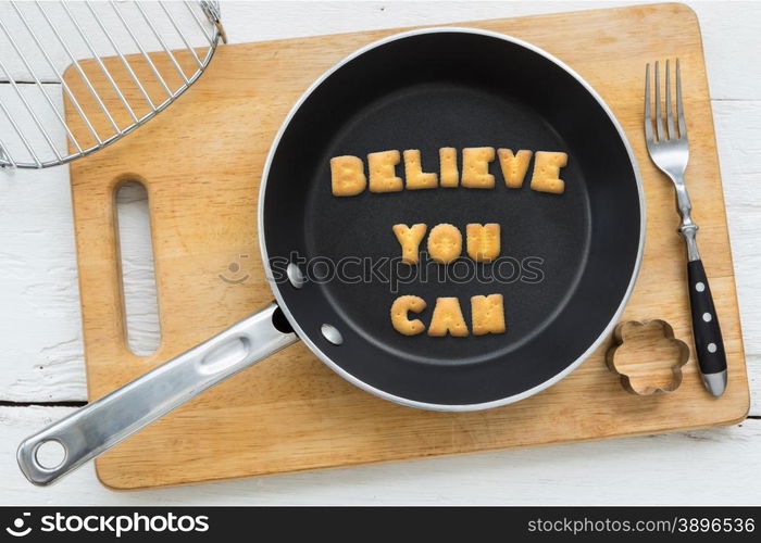 Top view of alphabet collage made of biscuits. Quote BELIEVE YOU CAN putting in black pan. Other kitchenware: fork, cookie cutter and chopping board putting on white wooden table, vintage style image.