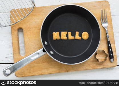 Top view of alphabet collage made of biscuits. Word HELLO putting in black pan. Other kitchenware: fork, cookie cutter and chopping board putting on white wooden table, vintage style image.