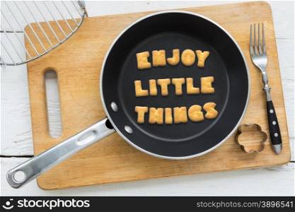 Top view of alphabet collage made of crackers. Quote ENJOY LITTLE THINGS putting in black pan. Other kitchenware: fork, cookie cutter and chopping board putting on white wooden table, vintage style image.