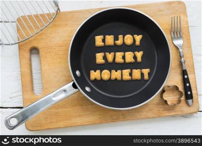 Top view of alphabet collage made of crackers. Quote ENJOY EVERY MOMENT putting in black pan. Other kitchenware: fork, cookie cutter and chopping board putting on white wooden table, vintage style image.