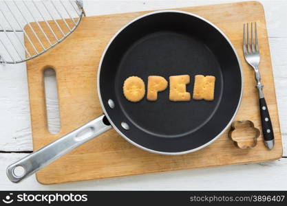 Top view of alphabet text collage made of cookies biscuits. Word OPEN putting in frying pan. Other utensils: fork, cookie cutter and cutting board putting on white wooden table, vintage style image.