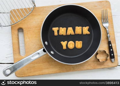 Top view of alphabet text collage made of cookies biscuits. Word THANK YOU putting in frying pan. Other utensils: fork, cookie cutter and cutting board putting on white wooden table, vintage style image.