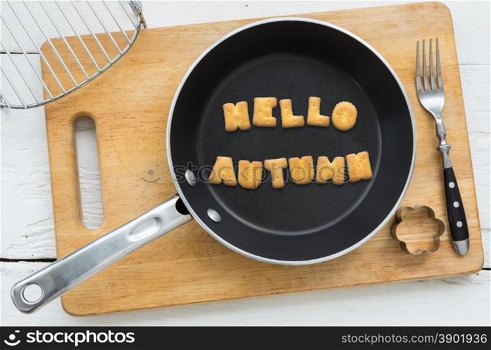 Top view of alphabet text collage made of cookies biscuits. Word HELLO AUTUMN in frying pan. Other utensils: fork, cookie cutter and cutting board putting on white wooden table, country and retro style.