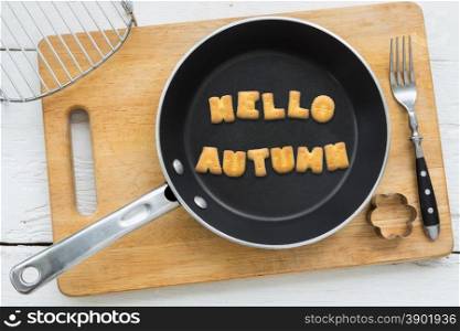 Top view of alphabet text collage made of cookies biscuits. Word HELLO AUTUMN in frying pan. Other utensils: fork, cookie cutter and cutting board putting on white wooden table, country and retro style.