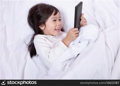 Top view of Asia little kid having fun playing smartphone lying on a bed in the morning on soft pillows laughing feels happy.