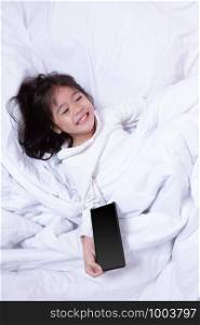Top view of Asia little kid having fun playing smartphone lying on a bed in the morning on soft pillows laughing feels happy.