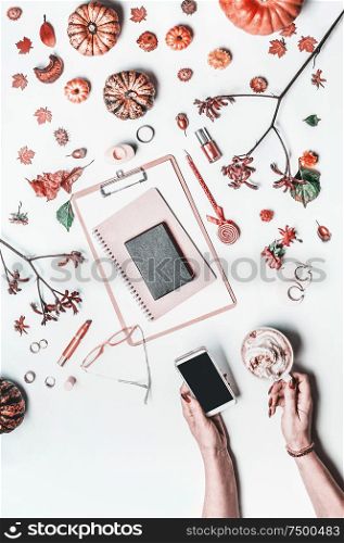 Top view of autumn home office table desk. Workspace with female woman hands holding smartphone and cup of hot chocolate , clipboard, notebooks, fall leaves and flowers, little pumpkins and cosmetics