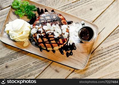 Top view of banana pancakes with macadamia nut and chocolate syrup topping on stack of pancakes and vanilla ice cream on wooden plate on wooden table.