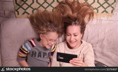 Top view of beautiful young woman and her lovely daughter doing selfie with smart phone and smiling on sofa at home. Two are lying on their backs with their amazing blonde long hair down on the floor.