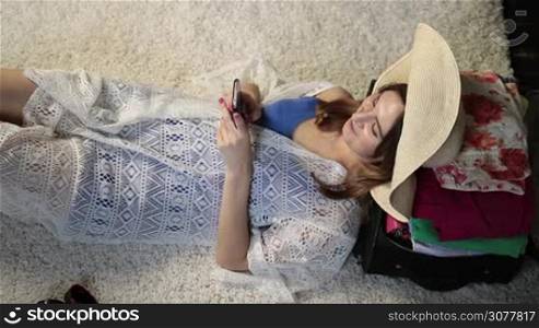 Top view of beautiful young woman in panama hat lying on the floor, her head on packed suitcase, typing message on mobile phone and smiling. Stunning female using cell phone after finishing packing luggage for vacation trip.