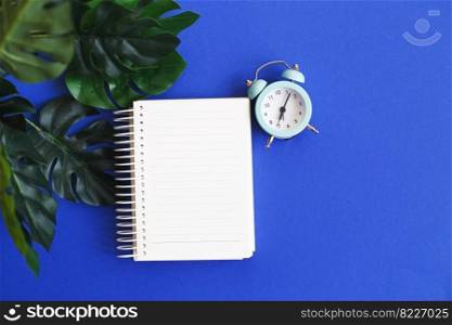 Top view of blank note paper, alarm clock and monstera leaf on blue background.. Top view of blank note paper, alarm clock and monstera leaf on blue background