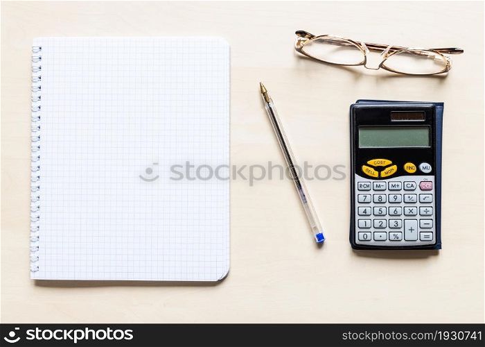 top view of blank sheet of squared paper of spiral notebook, pen, eyeglasses and financial calculator on light brown wooden board
